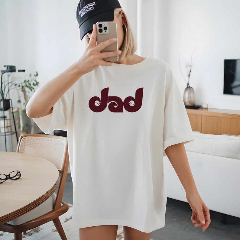 May include: Cream-colored t-shirt with the word "Dad" in a burgundy, retro-style font. The shirt has short sleeves and a relaxed fit. The person in the image is wearing a black baseball cap and holding a phone.