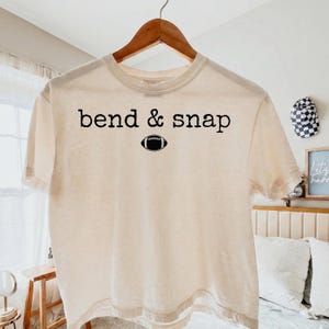 May include: A cream-colored short-sleeve t-shirt featuring the black text "bend & snap" printed across the chest, positioned above a small black graphic of an American football. The shirt is displayed on a wooden hanger against a neutral background.
