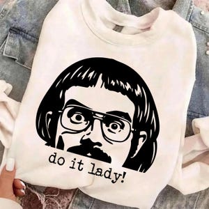 May include: White sweatshirt featuring a black and white graphic of a man with glasses and a mustache. The text "do it lady!" is printed below the graphic. The sweatshirt has a crew neck and long sleeves.