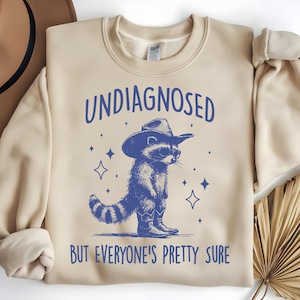 Puede incluir: Una sudadera beige con un mapache azul que lleva un sombrero de vaquero y gafas de sol. El texto "UNDIAGNOSED BUT EVERYONE'S PRETTY SURE" está impreso en la sudadera.