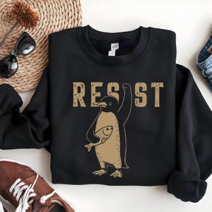 May include: Black sweatshirt featuring a brown penguin holding a fish and the word "RESIST" in brown text.