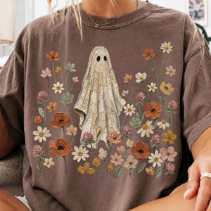 Peut inclure: Un t-shirt marron à manches courtes avec un motif central représentant un fantôme composé de pièces de tissu en patchwork. Le fantôme est entouré de fleurs de style brodé dans des tons orange, jaune, rose, blanc et vert.