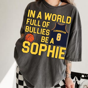 May include: A charcoal gray short-sleeve t-shirt featuring yellow text that reads IN A WORLD FULL OF BULLIES BE A SOPHIE. The design includes an orange basketball graphic and a navy blue basketball jersey with the name CUNNINGHAM and number 8.