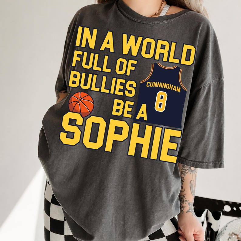 May include: A charcoal gray short-sleeve t-shirt featuring yellow text that reads IN A WORLD FULL OF BULLIES BE A SOPHIE. The design includes an orange basketball graphic and a navy blue basketball jersey with the name CUNNINGHAM and number 8.