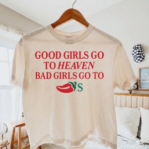 May include: A cream-coloured t-shirt with the text "GOOD GIRLS GO TO HEAVEN BAD GIRLS GO TO" in red. Below the text is a red chilli pepper graphic with the word "YS" in green. The shirt is hanging on a wooden hanger.