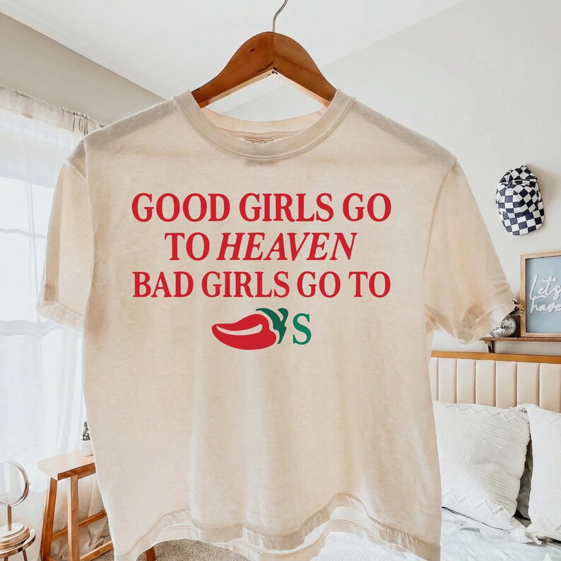 May include: A cream-coloured t-shirt with the text "GOOD GIRLS GO TO HEAVEN BAD GIRLS GO TO" in red. Below the text is a red chilli pepper graphic with the word "YS" in green. The shirt is hanging on a wooden hanger.