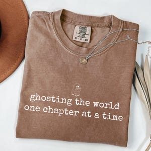 May include: A brown short-sleeve t-shirt with a crew neckline featuring a white graphic of a ghost reading a book above the text "ghosting the world one chapter at a time". The shirt has a Comfort Colors brand label inside the collar.