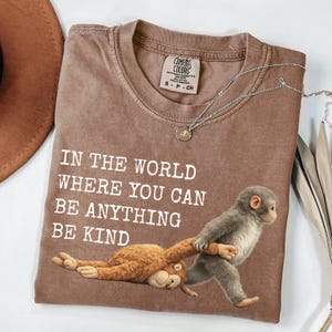 May include: A brown t-shirt featuring the text "IN THE WORLD WHERE YOU CAN BE ANYTHING BE KIND" and a monkey graphic. A silver necklace with a small pendant is placed on the shirt.