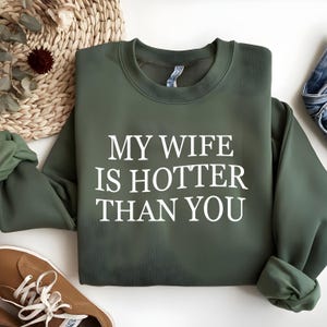 May include: An olive green sweatshirt with the text "MY WIFE IS HOTTER THAN YOU" in white. The sweatshirt is folded, with the sleeves and collar visible. Brown shoes and a woven basket are in the background.