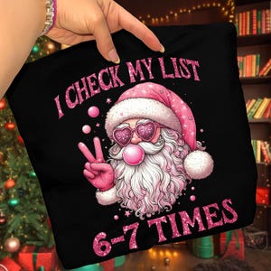 May include: Black cushion featuring a pink-clad Santa Claus with heart-shaped sunglasses, blowing a bubblegum bubble and giving the peace sign. Text reads "I CHECK MY LIST" and "6-7 TIMES" in pink glitter.