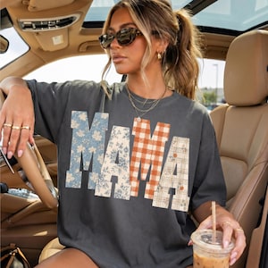 以下が含まれることがあります： 大きなブロック体で「MAMA」とプリントされたチャコールグレーの半袖Tシャツ。各文字は異なるパターンで、青い花柄、クリーム色の花柄、オレンジと白のギンガムチェック、小さなオレンジの花柄があしらわれたクリーム色のチェック柄が特徴です。