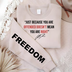 May include: A beige sweatshirt with the text "JUST BECAUSE YOU ARE OFFENDED DOESN'T MEAN YOU ARE RIGHT" in black and red. The sleeve displays the word "FREEDOM" in large black letters.