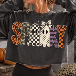 May include: A charcoal gray long-sleeve sweatshirt with a raw-edge hem. The front features the word SPOOKY in large letters. The S is orange leopard print, P is camouflage, O is black and white checkerboard, O is a ghost with a bow, K is purple stripes, and Y is orange and black checkerboard.