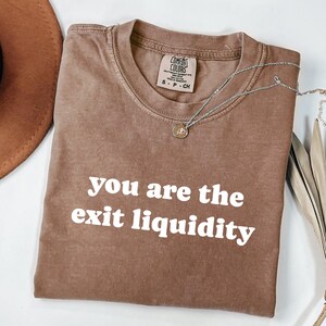 May include: A brown Comfort Colors t-shirt with the text "you are the exit liquidity" in white. A silver necklace with a small pendant is draped across the shirt. The shirt is size S-P-CH.