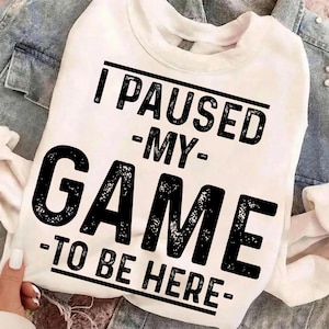 May include: White sweatshirt with the text "I PAUSED -MY- GAME -TO BE HERE-" in black, bold letters. The sweatshirt is held by a person, and is set against a denim jacket background.