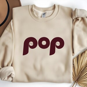 May include: A cream-colored sweatshirt with the word "pop" in a burgundy, retro-style font. The sweatshirt has a crew neck and long sleeves. The word "pop" is centered on the front of the shirt.