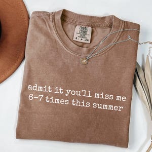 Op de afbeelding: Een bruin Comfort Colors t-shirt met de tekst "admit it you'll miss me 6-7 times this summer" in het wit. Een zilveren ketting met een kleine hanger ligt over het shirt. Een bruine hoed is zichtbaar linksboven.