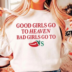 May include: White sweatshirt featuring the text "GOOD GIRLS GO TO HEAVEN BAD GIRLS GO TO" in red, with a red chili pepper graphic and the letter "S" in green. The sweatshirt is a casual style.