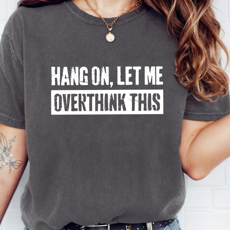 Op de afbeelding: Donkergrijs t-shirt met de witte tekst "HANG ON, LET ME OVERTHINK THIS". De tekst is vetgedrukt en zonder schreef, gecentreerd. Het t-shirt heeft korte mouwen en een ronde hals.