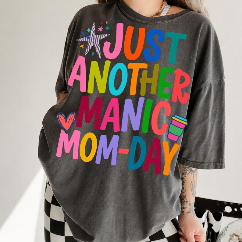 May include: Dark gray t-shirt with the text "JUST ANOTHER MANIC MOM-DAY" in colorful block letters. The design includes a star, heart, and coffee cup graphic. The shirt has a relaxed fit.