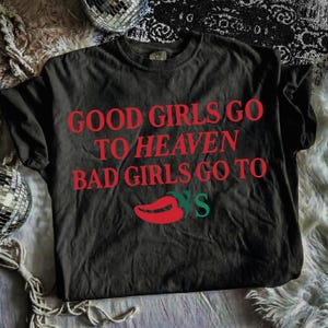 May include: Dark grey t-shirt featuring the red text "GOOD GIRLS GO TO HEAVEN BAD GIRLS GO TO" and a red chili pepper with green leaves. The shirt is a charcoal colour.