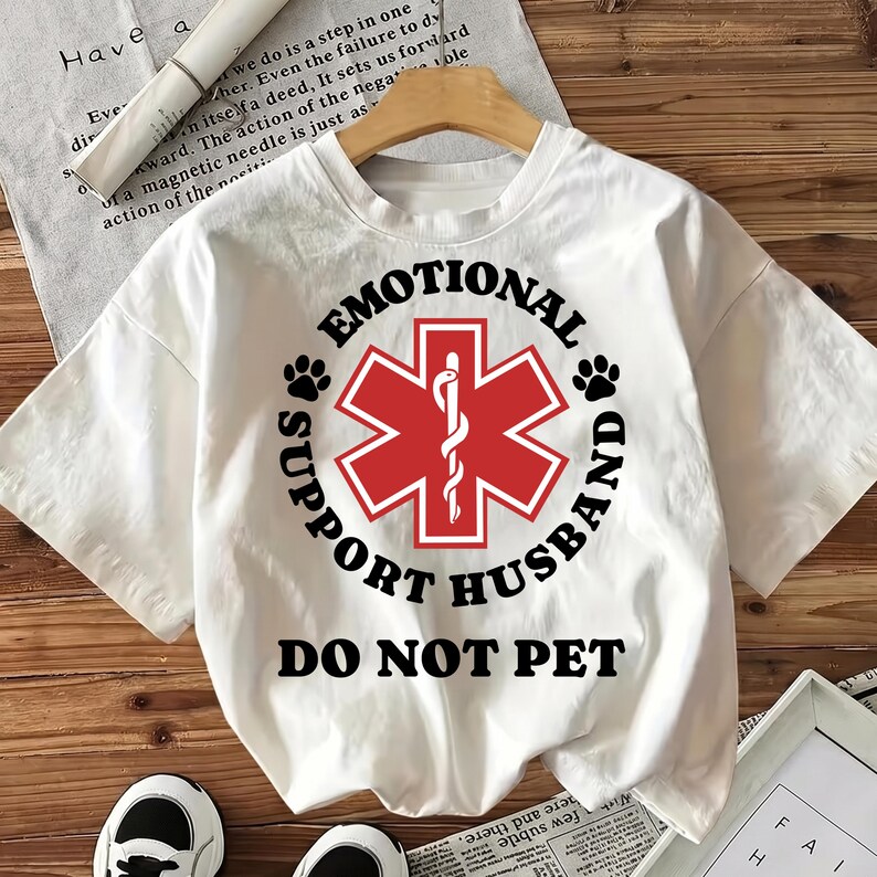 May include: White t-shirt with the text "EMOTIONAL SUPPORT HUSBAND DO NOT PET". The design includes a red medical symbol and paw prints. The shirt is made of a soft material.