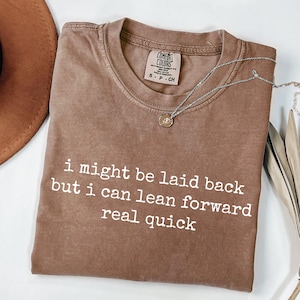 May include: A brown short-sleeve t-shirt with a crew neckline and a Comfort Colors brand tag. The shirt features the white printed text: i might be laid back but i can lean forward real quick. A silver-toned necklace with a small round pendant rests on the shirt.