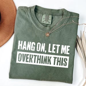 Op de afbeelding: Een olijfgroen t-shirt met de tekst "HANG ON, LET ME OVERTHINK THIS" in witte blokletters. Een gouden ketting met een kleine hanger ligt over het shirt. Het shirt is opgevouwen en ligt op een wit oppervlak.