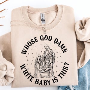May include: Beige sweatshirt featuring the text "WHOSE GOD DAMN WHITE BABY IS THIS?" in a circular design. The graphic shows a nativity scene with Mary, Joseph, and baby Jesus. A gold necklace is draped across the sweatshirt.