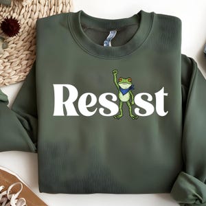 Puede incluir: Una sudadera verde oliva con la palabra "Resist" en letras blancas grandes. Una rana de dibujos animados con una bandana azul levanta el puño. La sudadera está hecha de un material suave.