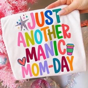 May include: White t-shirt with the text "JUST ANOTHER MANIC MOM-DAY" in colorful, playful lettering. The design includes a coffee cup graphic and a heart. The shirt is held up by a hand.
