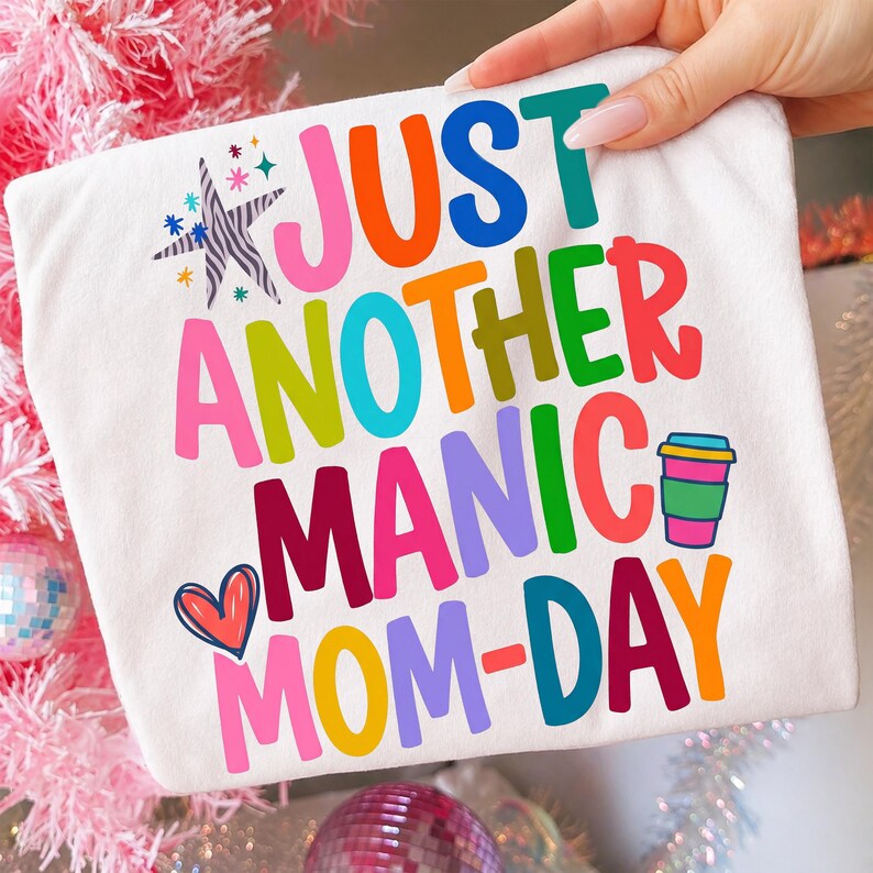 May include: White t-shirt with the text "JUST ANOTHER MANIC MOM-DAY" in colorful, playful lettering. The design includes a coffee cup graphic and a heart. The shirt is held up by a hand.