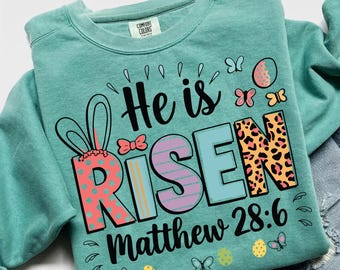 May include: A teal Comfort Colors sweatshirt with the words "He is Risen" in a colorful design. The word "Risen" has a leopard print pattern. The text "Matthew 28:6" is below. Easter-themed butterflies and eggs are also featured.