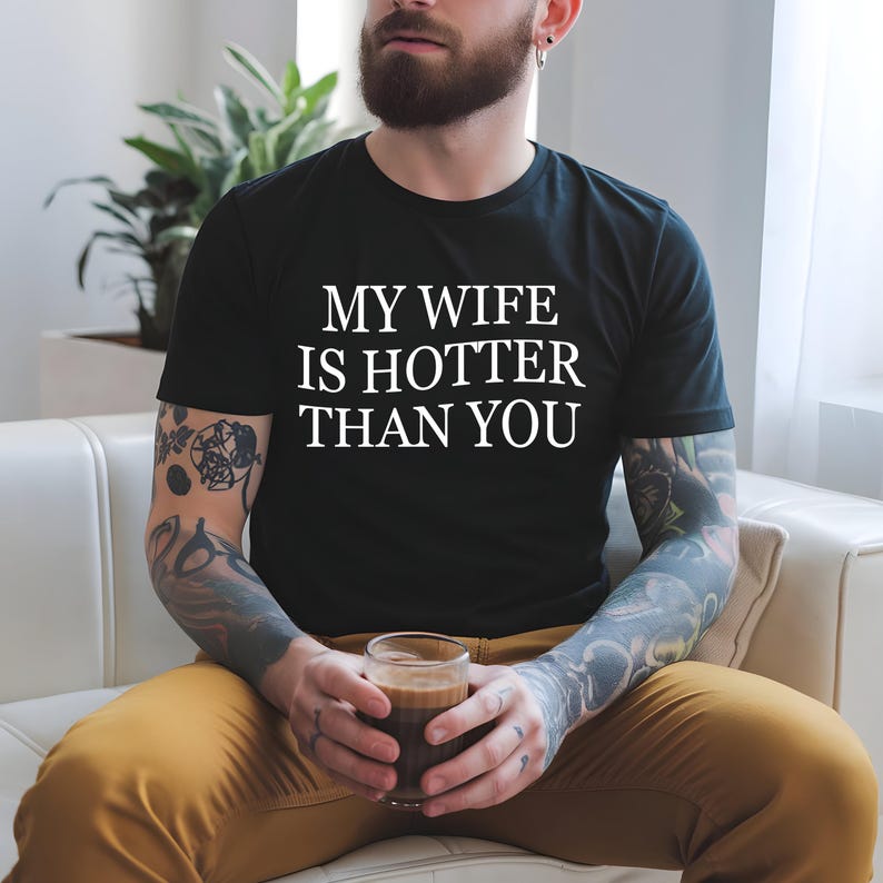 May include: Black t-shirt with the white text "MY WIFE IS HOTTER THAN YOU". The person is holding a glass of coffee. The person is wearing mustard-coloured trousers and has tattoos on their arms.