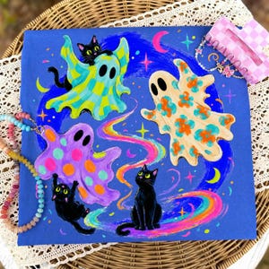 May include: A blue canvas painting featuring two black cats and three ghosts. One ghost is green and yellow checkered, one is beige with orange flowers, and one is purple with pink polka dots. A rainbow swirl and stars decorate the background.