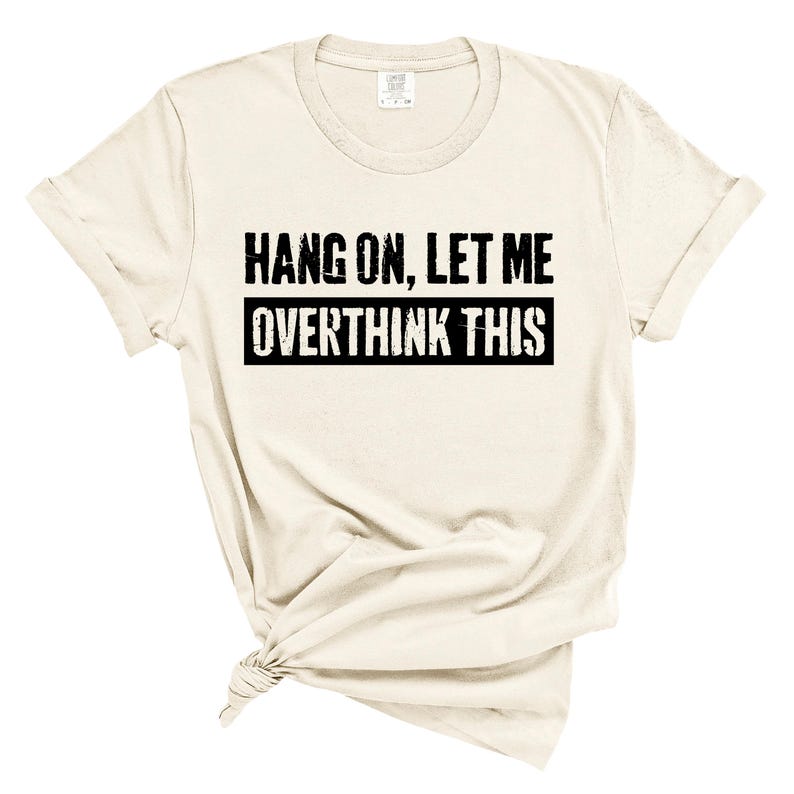 Op de afbeelding: Cr&egrave;mekleurig T-shirt met opgerolde mouwen. De tekst "HANG ON, LET ME OVERTHINK THIS" in zwarte, verweerde letters staat in het midden. Het shirt heeft een klassieke ronde hals en een relaxte pasvorm. De stof lijkt zacht.
