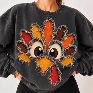 Puede incluir: Una sudadera de cuello redondo en color gris carbón con un gran gráfico de pavo en el centro. El pavo está formado por parches de tela superpuestos con bordes deshilachados en tonos rojo, naranja, amarillo y marrón, con dos ojos grandes.