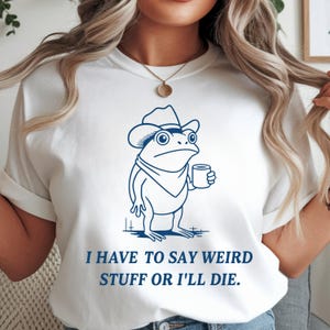Op de afbeelding: Wit T-shirt met een blauwe cartoon kikker met een cowboyhoed en een mok. De tekst "I HAVE TO SAY WEIRD STUFF OR I'LL DIE" staat in het blauw onder de kikkerillustratie.