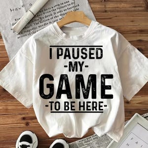 Pode incluir: Camiseta branca com o texto "I PAUSED MY GAME TO BE HERE" em letras pretas. A camiseta é exibida em um cabide de madeira. A camiseta é de manga curta e parece ser de algodão.