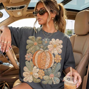 May include: A charcoal gray short-sleeve t-shirt featuring a large central graphic of a pumpkin surrounded by various floral shapes. The pumpkin and flowers are decorated with intricate, multi-colored patterns in shades of orange, green, and cream.
