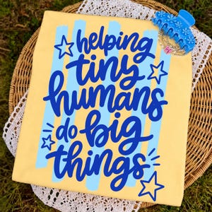 May include: A yellow t-shirt featuring vertical light blue brushstroke stripes and the dark blue text "helping tiny humans do big things" with star accents. A blue floral hair claw clip and a beaded bracelet are placed on the top right corner.