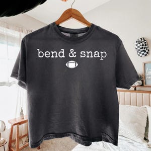 May include: A charcoal gray short-sleeve t-shirt featuring the white text "bend & snap" printed across the chest, positioned above a white graphic illustration of an American football. The shirt has a crew neckline and a relaxed fit.