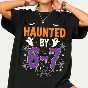 May include: Black t-shirt with the words "HAUNTED BY" in orange and white, and the numbers "6-7" in purple. The design includes white ghosts, bats, spiderwebs, and stars, perfect for Halloween.