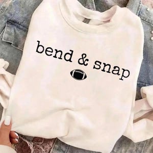 May include: A white long-sleeve sweatshirt featuring the black text "bend & snap" printed across the chest. Below the text is a small, black and white graphic illustration of an American football. The fabric appears soft and casual in style.