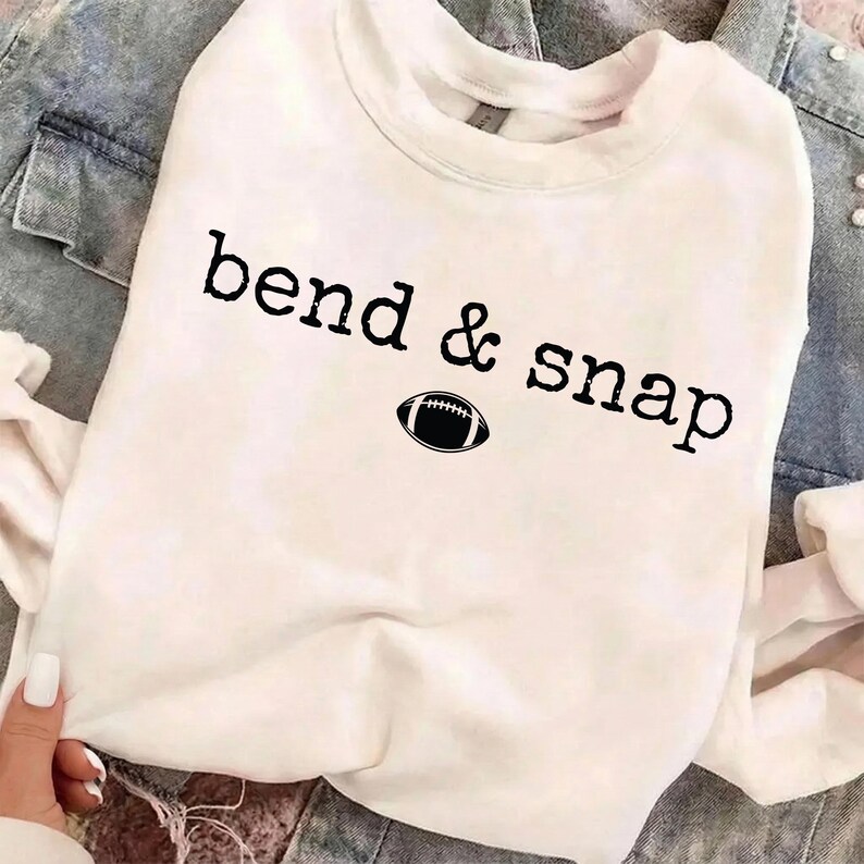 May include: A white long-sleeve sweatshirt featuring the black text "bend & snap" printed across the chest. Below the text is a small, black and white graphic illustration of an American football. The fabric appears soft and casual in style.