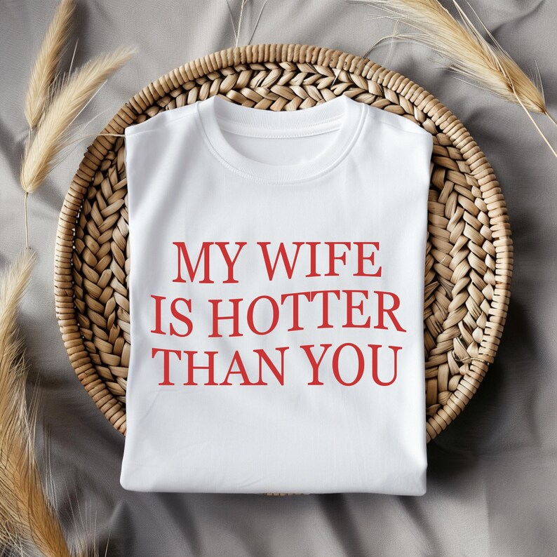 May include: White t-shirt with the red text "MY WIFE IS HOTTER THAN YOU." The shirt is folded on a round, woven wicker tray. Dried wheat stalks are placed on the side of the tray.