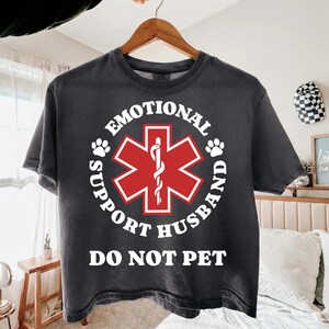 May include: Dark gray t-shirt with the words "EMOTIONAL SUPPORT HUSBAND DO NOT PET" in white. The design includes a red and white medical symbol and paw prints. The shirt is hanging on a wooden hanger.