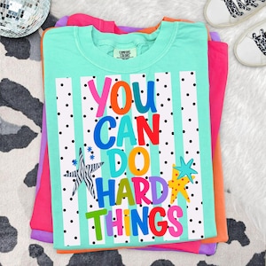 May include: A mint green Comfort Colors t-shirt with the text "YOU CAN DO HARD THINGS" in colorful letters. The design includes black polka dots and star graphics. The shirt is stacked on top of other colorful shirts.