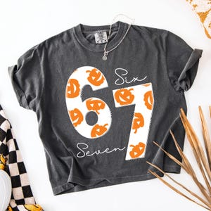 May include: Dark gray t-shirt with the numbers "67" in white, filled with orange jack-o'-lanterns. The word "Six" is above the "6" and "Seven" is below the "7", both in white script. A silver necklace is draped on the shirt.