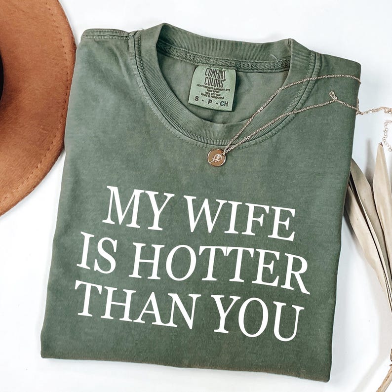 May include: An olive green t-shirt with the white text "MY WIFE IS HOTTER THAN YOU". A gold-coloured necklace with a pendant lies on the folded shirt. The shirt is on a white surface. The t-shirt has a Comfort Colors label.